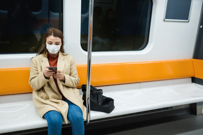 Woman wearing mask using mobile phone while sitting at train