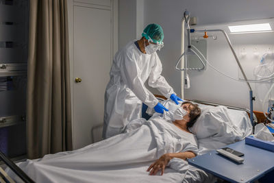 Male professional practitioner in medical uniform and protective mask and gloves visiting and caring elderly female patient lying in bed in mask in hospital ward while curing coronavirus