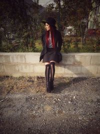 Full length of woman wearing top hat while sitting on retaining wall