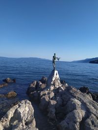 Man standing on rocks by sea against clear blue sky