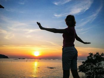 Rear view of woman with arms raised standing at sunset