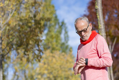 Senior man in sports clothing checking time on smart watch while leaning on tree