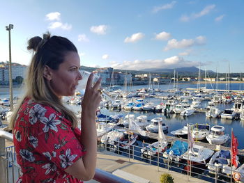 Side view of woman drinking wine while standing against boats moored at harbor