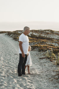 Father and preschool aged girl standing in sand at beach looking away