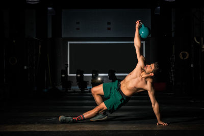 Muscular male athlete in shorts doing side plank and lifting kettlebell during functional workout in dark gym