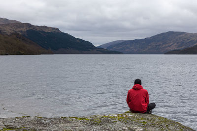 Rear view of man sitting on rock by river against sky