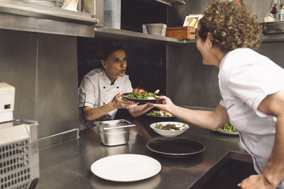 Female chef talking to coworker while giving food plate in commercial kitchen