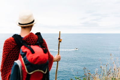 Back view of anonymous female hiker with backpack and stick standing on hill and admiring rippling sea with floating boat while resting during camino de santiago route in spain