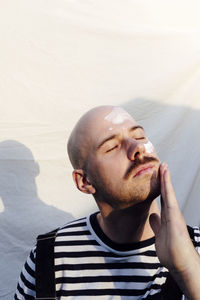 Bald man with eyes closed applying moisturizer in front of white backdrop