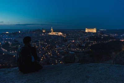 Back view of silhouette of male traveler sitting on hill and enjoying spectacular night cityscape of old city toledo in spain with illuminated medieval buildings