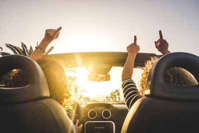 Rear view of friends with arms raised sitting in car against clear sky during sunset
