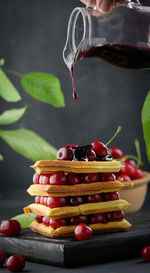 A stack of baked belgian waffles with ripe red cherries, a hand pours jam from a transparent jug