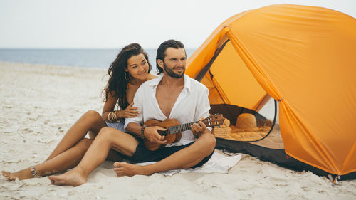 Full length of couple sitting on beach by tent
