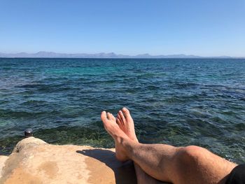 Low section of man relaxing on rock by sea against sky