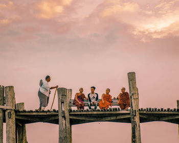 People sitting on railing against sky during sunset