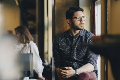 Thoughtful businessman holding smart phone while looking through window at office