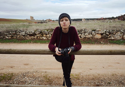 Portrait of young woman with camera standing by railing against cloudy sky