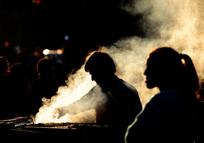 Silhouette man standing with people while preparing food on barbecue grill amidst smoke