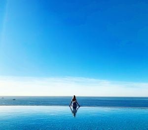 Rear view of young woman standing in infinity pool against blue sky