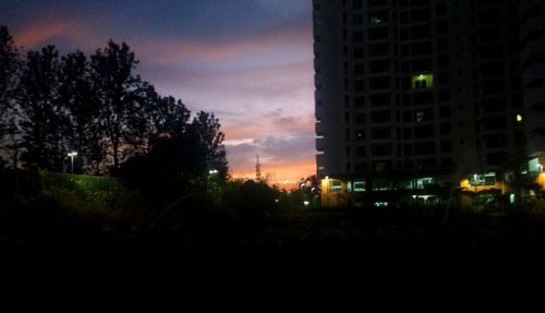 Silhouette trees by illuminated city against sky at sunset