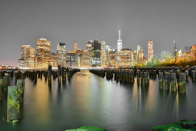 Wooden posts on hudson river by illuminated city skyline against clear sky at dusk