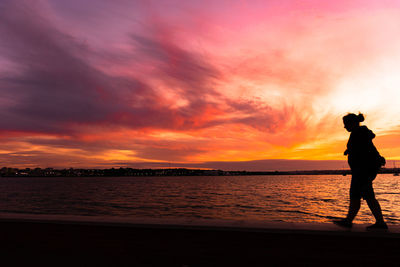 Silhouette woman walking on retaining wall by sea against orange sky