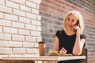 Young woman using mobile phone while sitting against brick wall