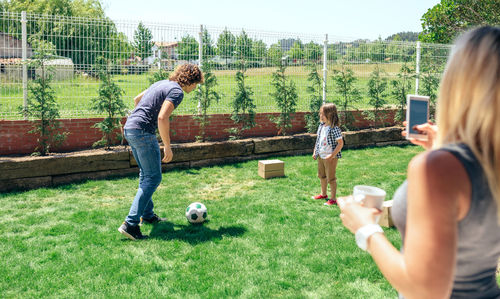 Mother using phone while father and son playing soccer in lawn