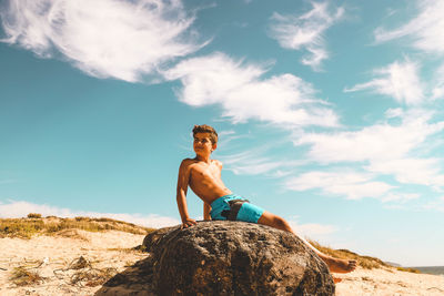 Young man sitting on rock against sky