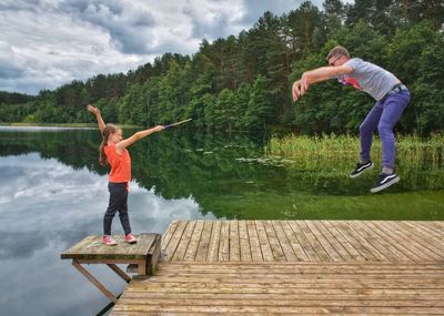 Full length of man jumping with girl holding stick on pier over lake against sky