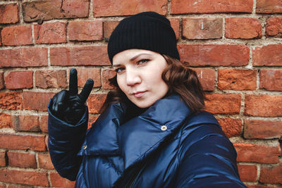 Portrait of young woman gesturing peace sign while standing against brick wall
