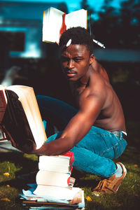 Shirtless man reading book while crouching on field