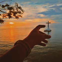 Close-up of human hand pointing at religious cross in sea against sky at sunset