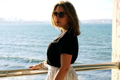 Young woman wearing sunglasses standing by sea against sky