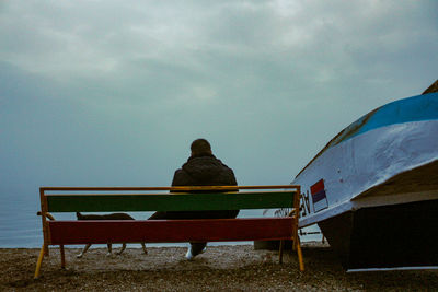 Rear view of man sitting on bench at beach against sky