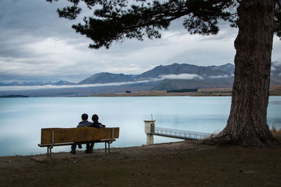 Rear view of man sitting on bench by lake against sky