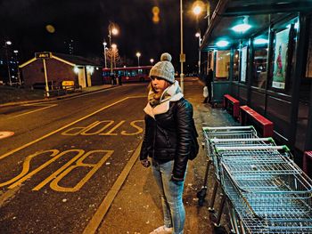 Young woman in warm clothing standing on sidewalk at night