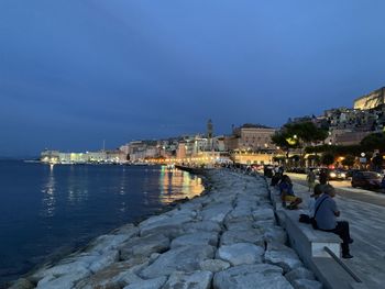 Illuminated buildings by sea against blue sky at dusk