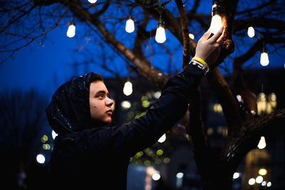 Low angle view of man holding illuminated light bulb hanging from tree at night