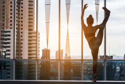 Rear view of young woman exercising against window in city during sunset