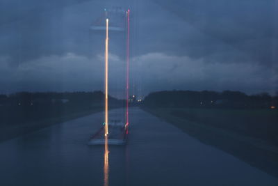 Illuminated road by lake against sky at dusk