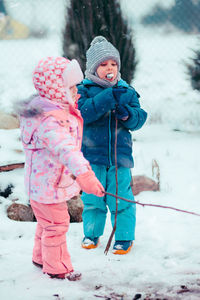 Full length of cute siblings eating marshmallow while standing on snow covered field