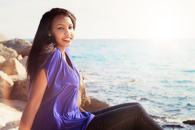 Portrait of smiling young woman sitting at sea against sky