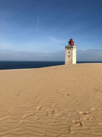 Lighthouse on sand dune by sea against sky