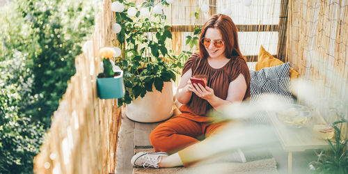 Woman wearing brown blouse and sunglasses works on modern phone siting on pillows on light terrace.