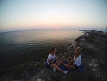 Women sitting on shore against sky during sunset