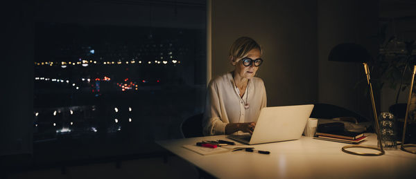 Mature female professional working late while using laptop at illuminated desk in office