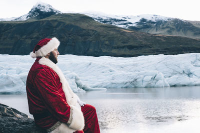 Side view of man wearing santa claus costume looking at lake during winter