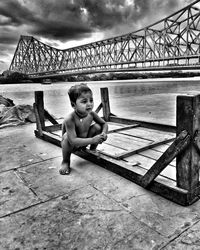 Portrait of shirtless boy sitting on bridge against sky