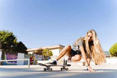 Confident young blond woman skateboarding at park against clear blue sky on sunny day